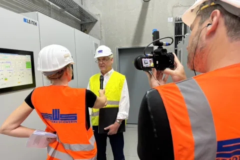 Zwei Personen in orangenen Warnwesten mit Stadtwerke Logo interviewen einen Herren vor einem Schaltschrank.