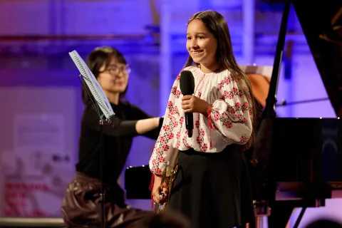 Junge Musikerin hält eine Ansprache mit Mikrophon, um Hintergrund sitzt ihre Lehrerin am Klavier