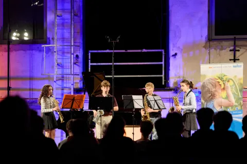 Vier junge Musiktalente mit Blasinstrumenten stehen auf einer blau beleuchteten Bühne