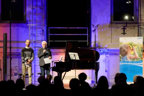 Zwei Herren stehen am linken Rand einer bunt ausgeleuchteten Bühne mit Piano.