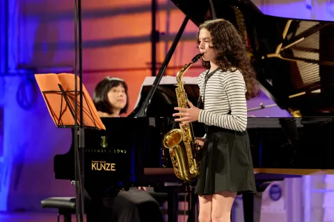 Junge Musikerin mit Saxophon wird im Hintergrund von ihrer Lehrerin am Klavier begleitet