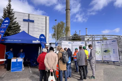 Vor dem Heizkraft stehen ein Pavillion der Stadtwerke sowie eine Ausstellung mit Informationen zur Unternehmnesgeschichte und eine Gruppe von Menschen, die sich für eine Führung bereit machen.