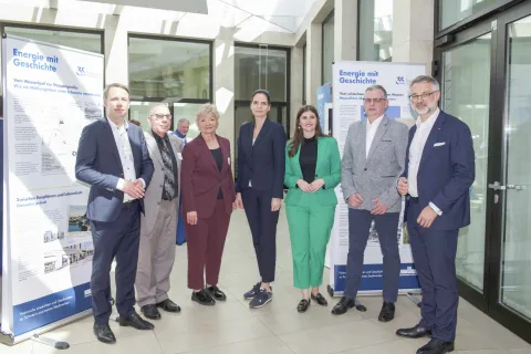 Sieben Personen im Businesslook stehen nebeneinander in einem sonnigen Foyer.