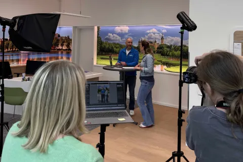 Im Hintergrund sind zwei Menschen in einer Beratungsszene. Im Vordergrund sieht man die Fotografin sowie eine Frau von Hinten beim Aufnehmen der Fotos und Sichtung am Laptop.