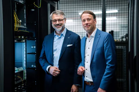 Zwei Geschäftsführer stehen vor einem Technikschrank mit Glasfaserkabeln.