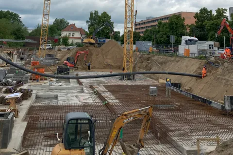 Baugrube mit Bagger und zwei Kränen, welche eine Fernwärmeleitung in der Luft halten.