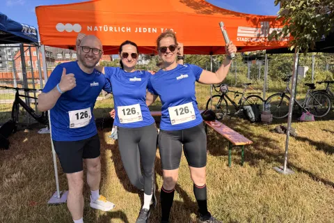 Ein Mann und zwei Frauen in Sportsachen mit blauem T-Shirt der Stadtwerke stehen vor dem Unternehmenspavillon.