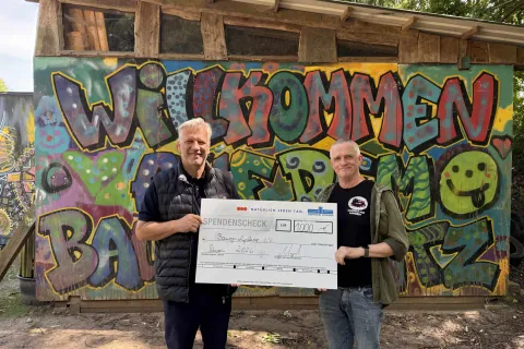 Zwei Männer stehen vor einer Graffitiwand des Bauspielplatzes und halten einen riesigen Spendencheck in der Hand.