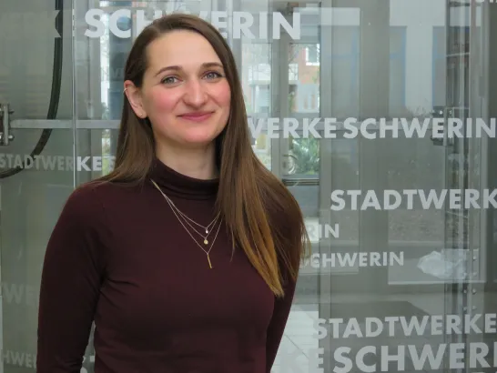 Frau mit langen braunen Haaren steht vor einer Glaswand mit mehreren Schriftzügen in weiß der Stadtwerke Schwerin
