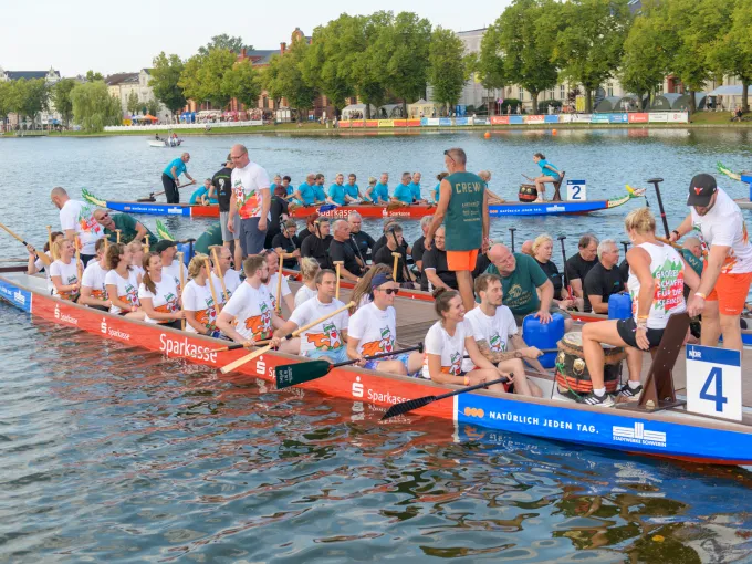 drei vollbesetzte Drachenboote legen vom Steg ab, die Mannschaften tragen einheitliche Teamshirts, Team im Vordergrund trägt weiße T-Shirts und ist euphorisch