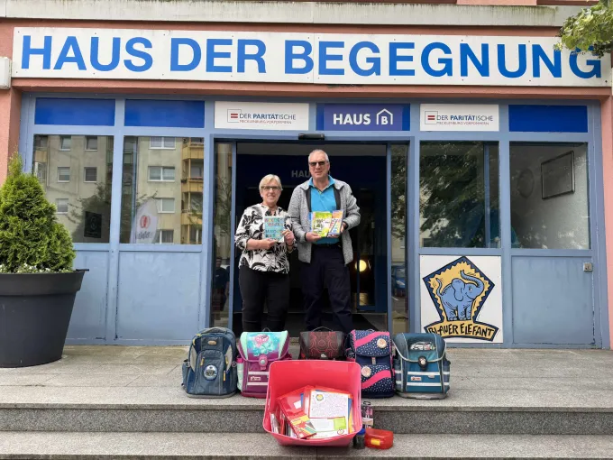 Zwei Personen stehen vor einem Haus und halten Schreibhefte in den Händen. Davor stehen Schulranzen und andere Schulmaterialien.