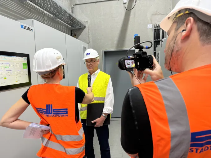 Zwei Personen in orangenen Warnwesten mit Stadtwerke Logo interviewen einen Herren vor einem Schaltschrank.
