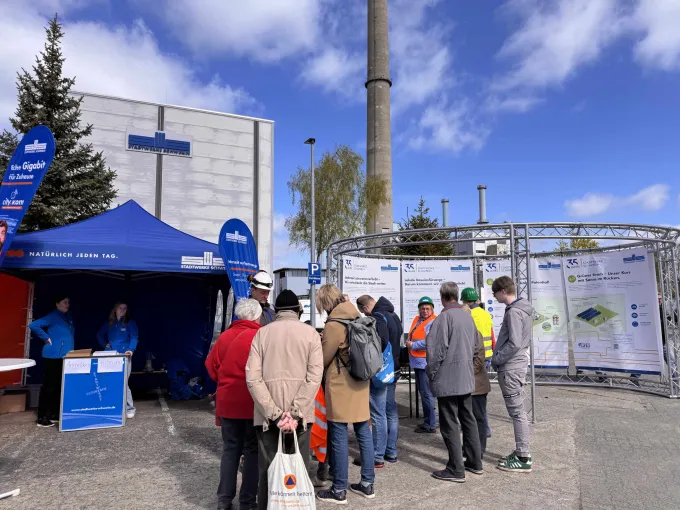 Vor dem Heizkraft stehen ein Pavillion der Stadtwerke sowie eine Ausstellung mit Informationen zur Unternehmnesgeschichte und eine Gruppe von Menschen, die sich für eine Führung bereit machen.