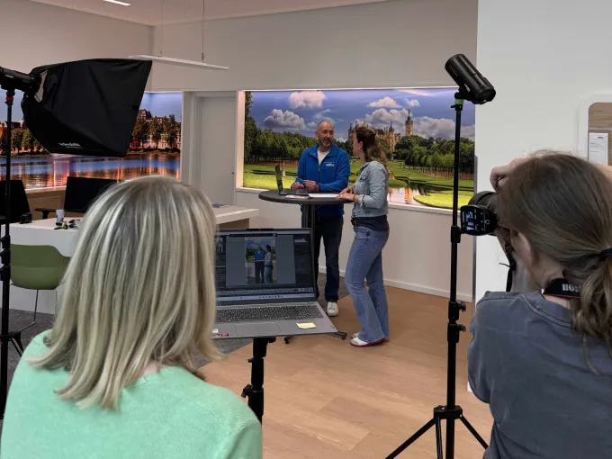 Im Hintergrund sind zwei Menschen in einer Beratungsszene. Im Vordergrund sieht man die Fotografin sowie eine Frau von Hinten beim Aufnehmen der Fotos und Sichtung am Laptop.