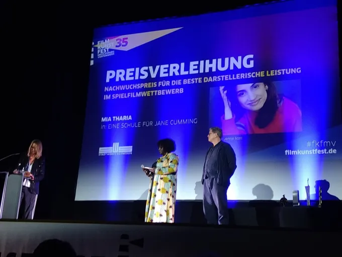 Aufnahme der Bühne beim Filmkunstfest MV mit Moderatorin. Der Screen zeigt die Gewinnerin des Nachwuchsdarstellerpreises.