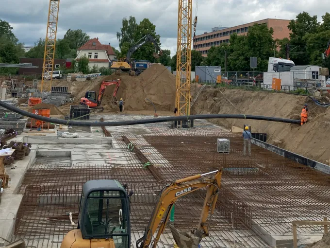 Baugrube mit Bagger und zwei Kränen, welche eine Fernwärmeleitung in der Luft halten.