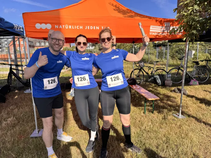 Ein Mann und zwei Frauen in Sportsachen mit blauem T-Shirt der Stadtwerke stehen vor dem Unternehmenspavillon.