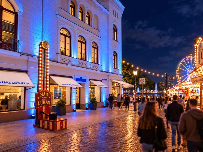 Das Kundencenter der Stadtwerke in der Mecklenburgstraße ist mit blauem Licht angestrahlt, davor steht ein "Hau den Lukas". Auf der anderen Straßenseite stehen jahrmarktstypische Imbissbuden.