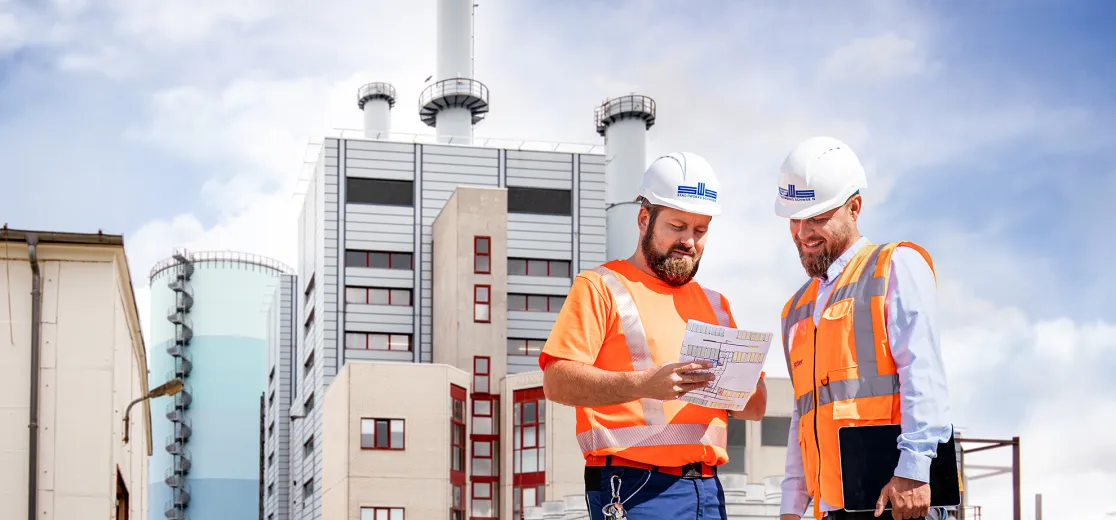 Zwei Männer mit Warnweste und Helm stehen vor dem Heizkraftwerk Schwerin Süd und schauen auf einen Plan.