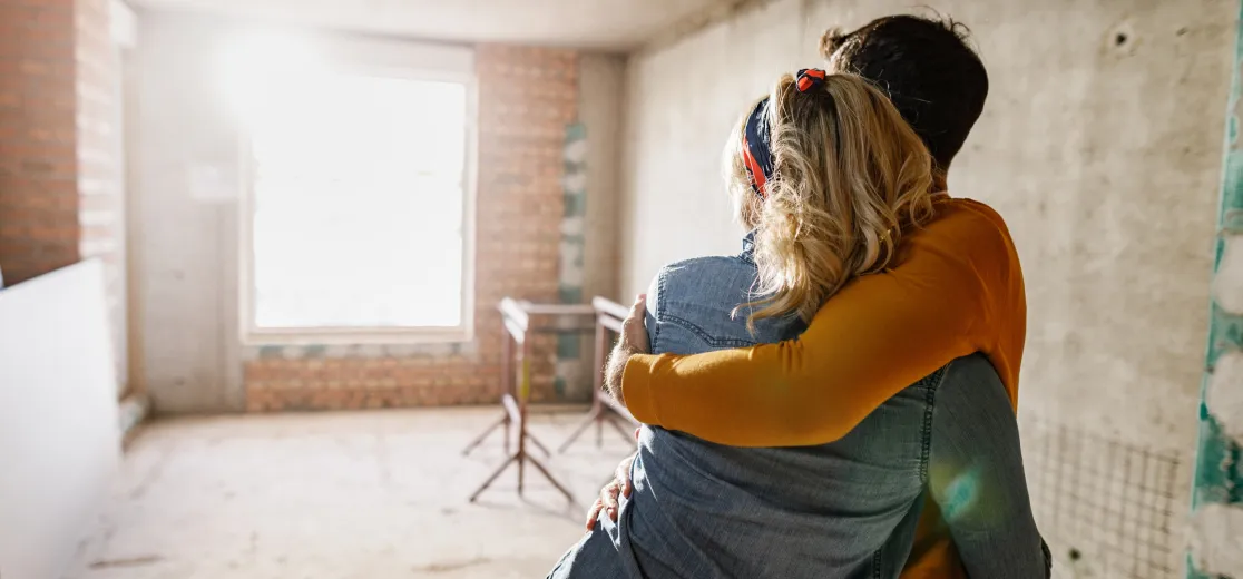 Ein junges Paar steht im Rohbau seines neuen Zuhauses und hält sich glücklich in den Armen.