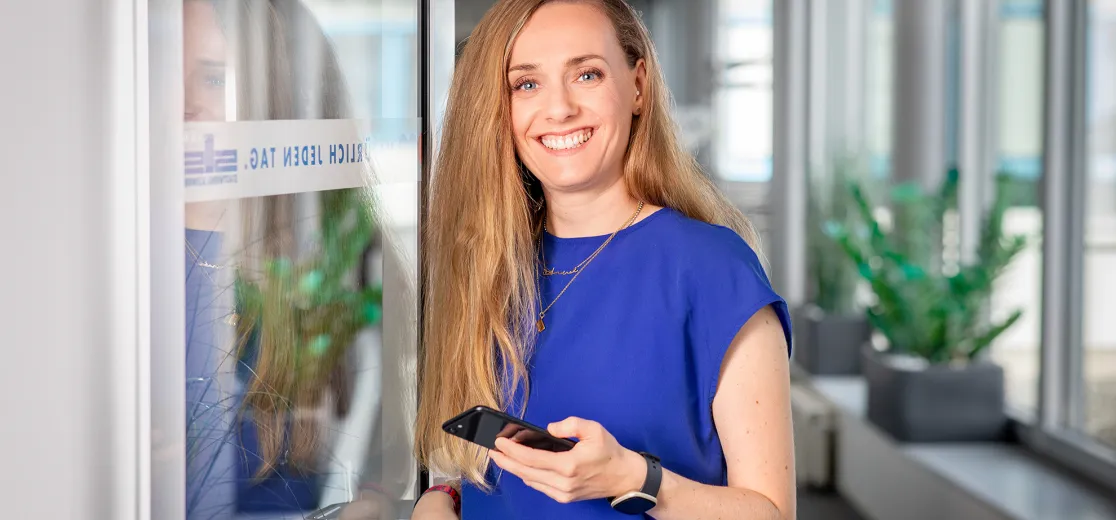 Eine junge Frau steht lächelnd in einer offenen Tür. Sie hält ein Smartphone in der Hand.