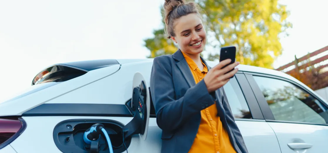 Junge Frau lehnt mit dem Handy in der Hand am Auto, während dieses lädt.