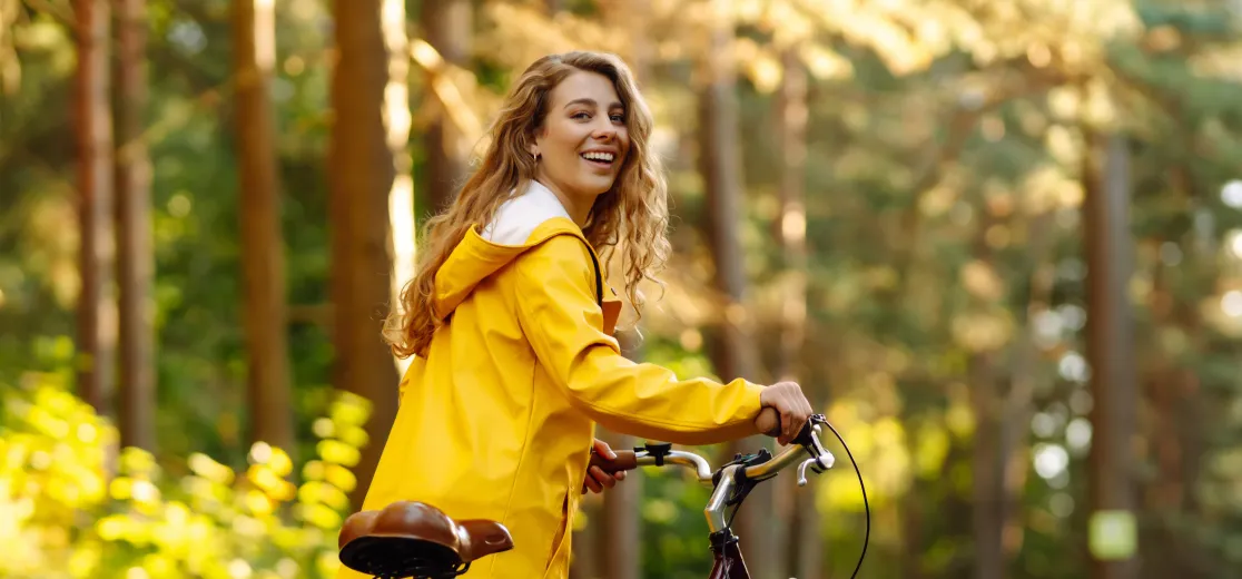 Eine Frau im gelben Mantel fährt mit Ihrem Fahrrad durch einen Wald.