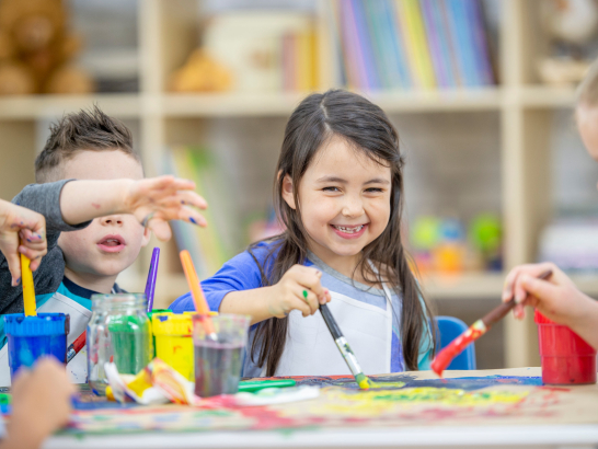 Nahaufnahme von drei Kindern im Vorschulalter beim gemeinsamen Malen mit verschiedenen Farben, Mädchen mit langen schwarzen Haaren steht im Mittelpunkt und schaut in die Kamera