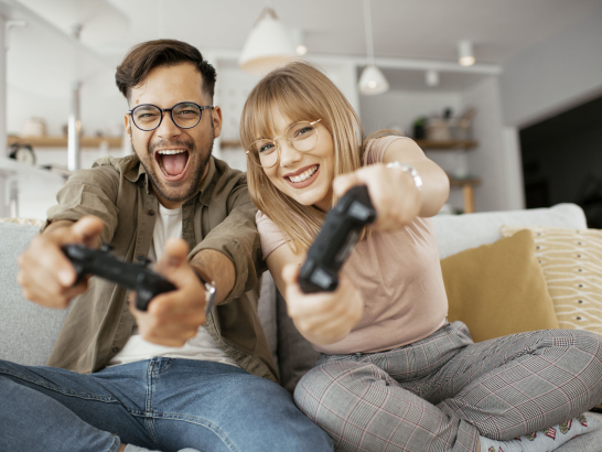 Ein Mann und eine Frau sitzen mit Controllern auf der Couch und gucken fröhlich in die Kamera.