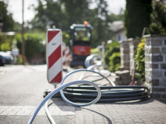 Leerrohre für Glasfaser liegen neben einer Baustellenbake auf einem Gehweg mit Minibagger im Hintergrund.