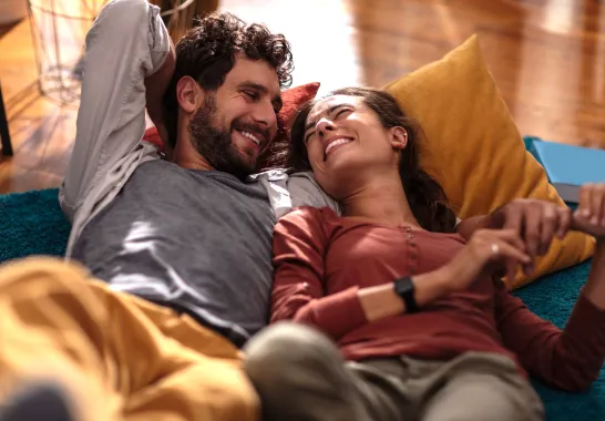 Ein junges Paar liegt gemeinsam auf dem Sofa und lächelt sich an.