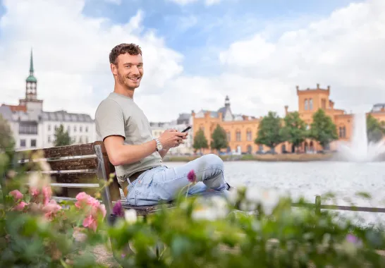 Ein Mann sitzt auf einer Bank vor dem Schweriner Pfaffenteich und surft auf dem Handy.