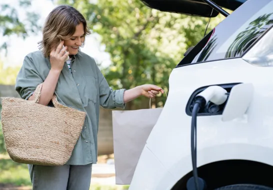 Eine Frau steht mit einer Einkaufstasche neben ihrem E-Auto, das gerade lädt.