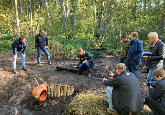 Acht Männer gruppieren sich um ein Stauwerk im Wald, in der Mitte kniet ein Landesforst Mitarbeiter und erklärt.