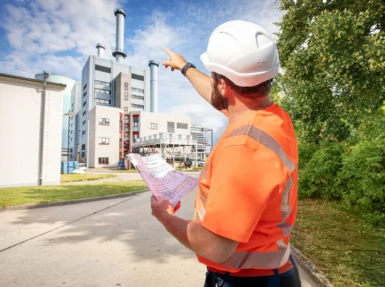 Ein Mann mit Warnweste und Helm zeigt mit dem Finger auf das Heizkraftwerk Schwerin Süd.