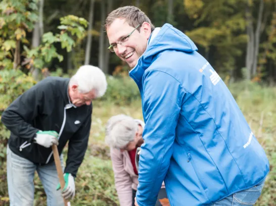 Ein Stadtwerke-Mitarbeiter und zwei weitere Personen pflanzen im Wald Baumsetzlinge ein.
