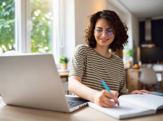 Junge Frau sitzt vor ihrem Laptop am Schreibtisch. Sie schreibt etwas auf einen Zettel.