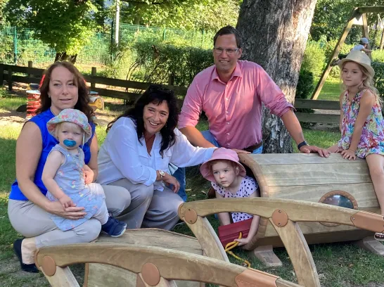 Zwei Frauen und ein Mann hocken bei Outdoor-Bauelementen aus Holz mit drei spielenden Kindern.