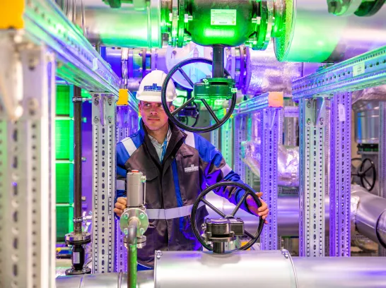 Ein Mitarbeiter der Stadtwerke Schwerin steht mit Arbeitskleidung und Helm hinter einem Rohrsystem mit großen Handrädern.