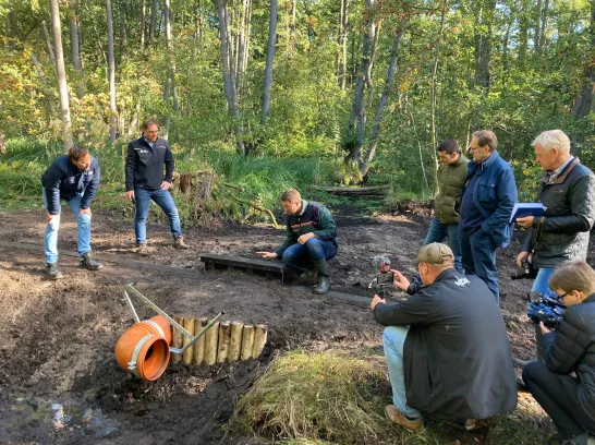 Acht Männer gruppieren sich um ein Stauwerk im Wald, in der Mitte kniet ein Landesforst Mitarbeiter und erklärt.