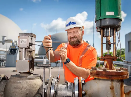 Mitarbeiter mit weißem Helm und Stadtwerkelogo vor den Kuppeln der Biogasanlage.