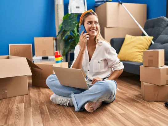 Junge Frau mit Laptop und Handy zwischen Umzugskartons.